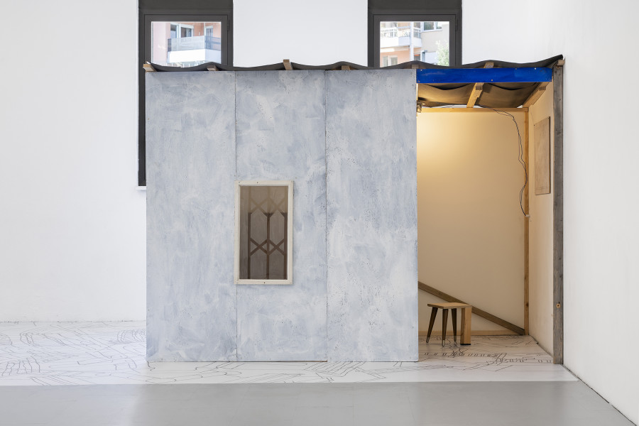 Aldo Mozzini, Quasi un rifugio, 2025.Drawing, large poster on poster paper, 356 x 504 cm – MDF panels, window with design made with yoghurt, bituminous paper, white wooden flag, stool, lamp, intaglio printing fabrics, plastic waste, various types of wood, 220 x 280 x 140 cm. Ph by Riccardo Giancola. Images courtesy of artists and La Rada, Photography by Riccardo Giancola (@riccardo_giancola) and Emanuele Finardi (@e.m.hey)